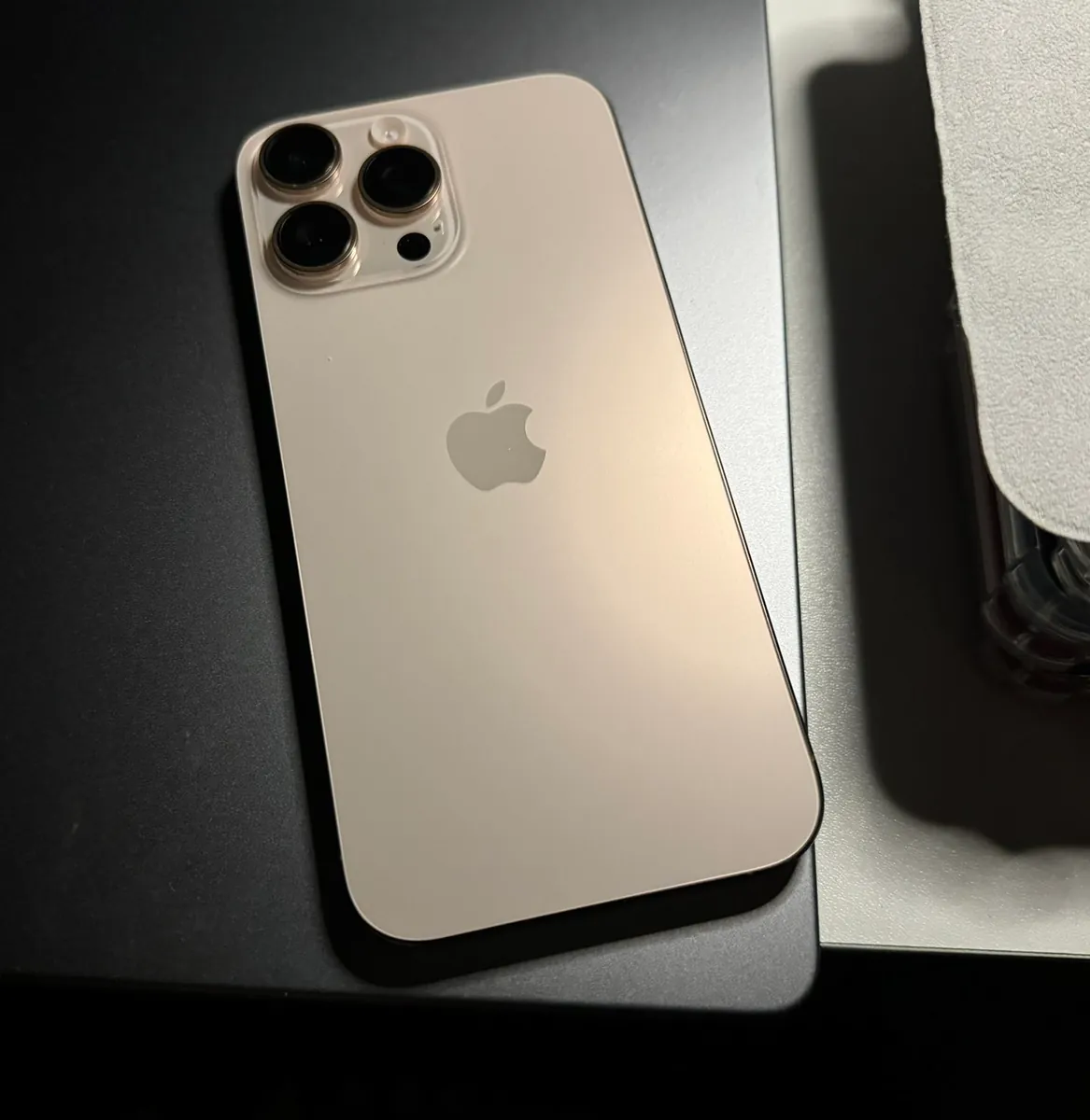 iPhone 16 Pro Max in Desert Titanium, back view, lying on a dark desk